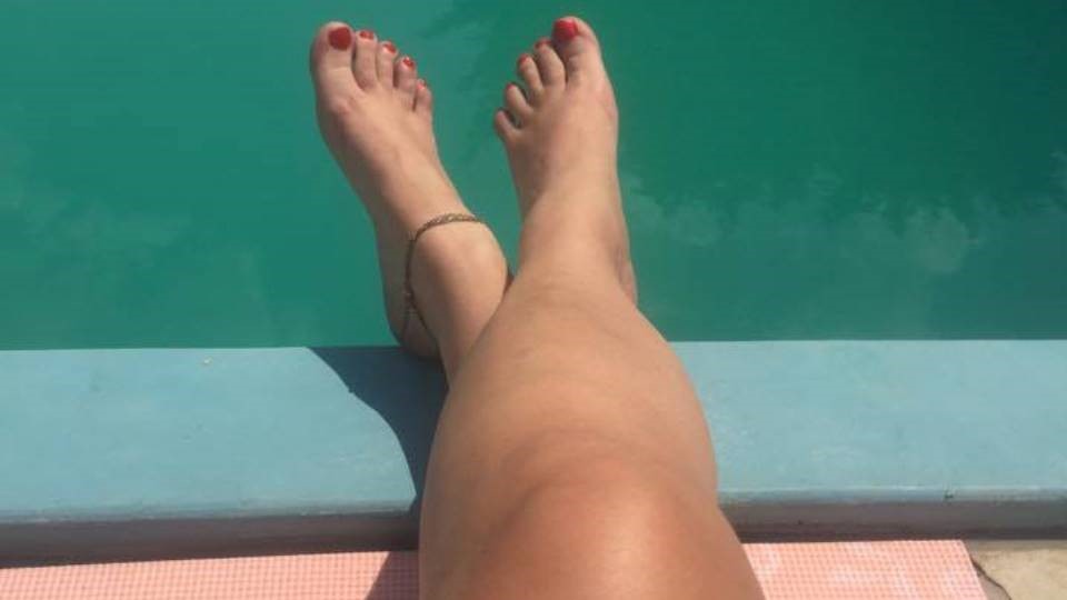 A képen két keresztbetett női lábfej látható egy medence partján, vörösre festett körmökkel, háttérben a medence vizével.